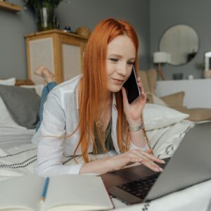 Red-haired woman multitasking in a cozy bedroom, using a laptop and phone for work.