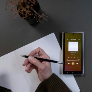 A cozy workspace scene with a hand taking notes, a smartphone playing a podcast, and a minimalist vase.